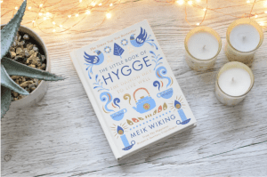A book laying on a table surrounded by cozy elements light candles and string lights. Danish Secrets to Hygge.